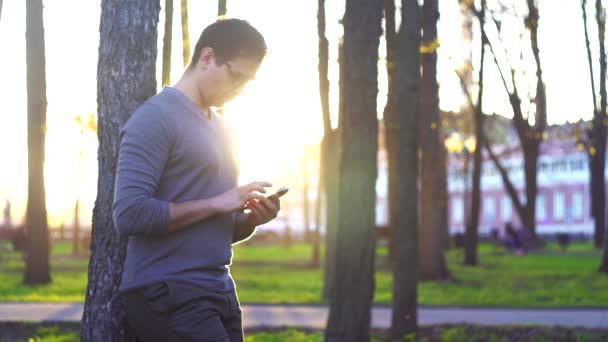 Pria di taman saat matahari terbenam terhadap latar belakang pemandangan kota menggunakan smartphone — Stok Video