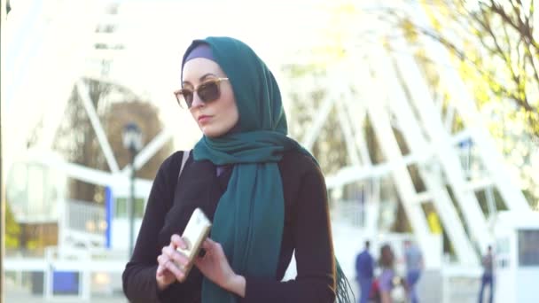 Дівчина в хіджабі і сонцезахисних окулярах чекає когось, дивлячись на годинник — стокове відео
