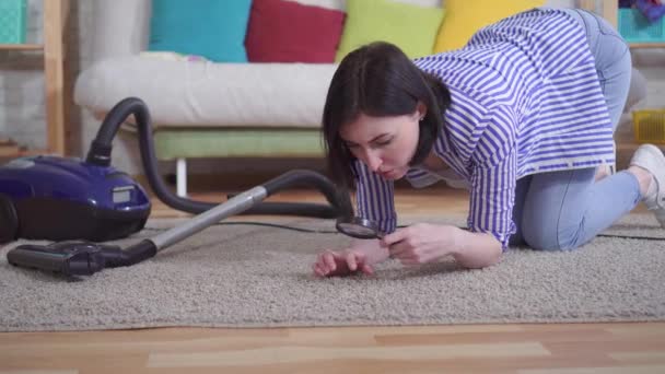 Junge Frau im Wohnzimmer untersucht den Teppich durch ein Vergrößerungsglas — Stockvideo