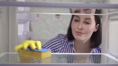 Eldivenli genç kadın buzdolabını yıkıyor.
