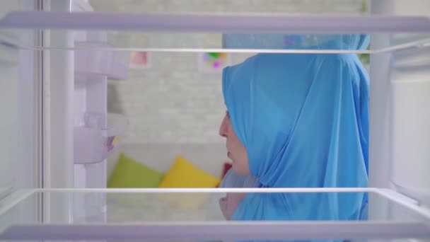 Mujer musulmana joven en un pañuelo nacional mira en un refrigerador vacío y pide comida en casa a través de una aplicación en su teléfono — Vídeos de Stock