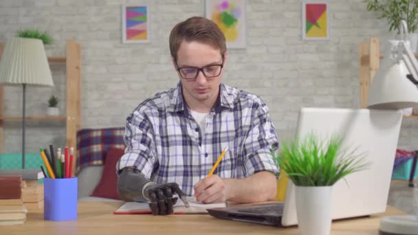 Young man with a bionic prosthetic hand at work uses a laptop and makes notes in a notebook — Stock Video