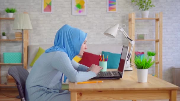 Young Muslim woman in a traditional headscarf slouches while sitting at a laptop and feels pain in her neck — Stock Video