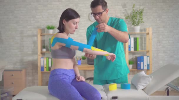 Фізіотерапевт у формі пристібає до рук пацієнта спеціальну кінезотапу. — стокове відео