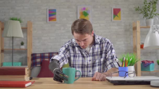 Молодий чоловік, який сидить за столом, бере чашку з біонічною протезом руки. — стокове відео