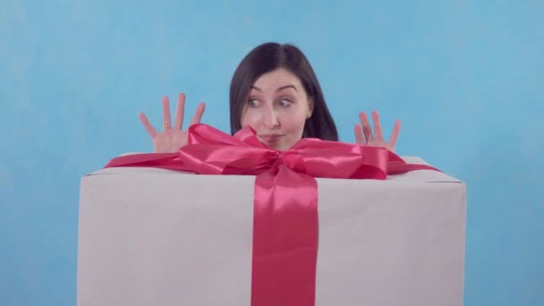 Divertida joven mujer mira hacia fuera desde detrás de un gran regalo empaquetado sobre un fondo azul — Vídeo de stock
