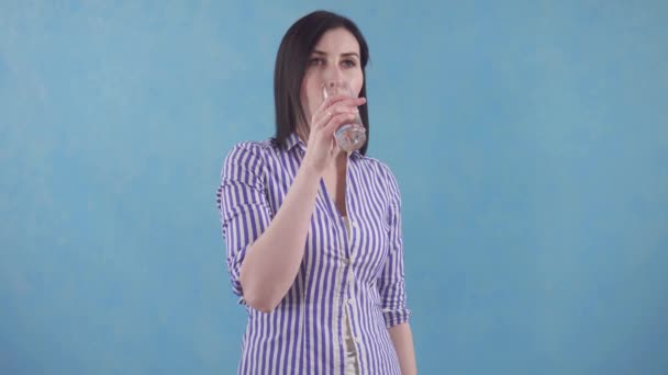 Young woman with bewilderment drinks water from a glass standing on a blue background — Stock Video