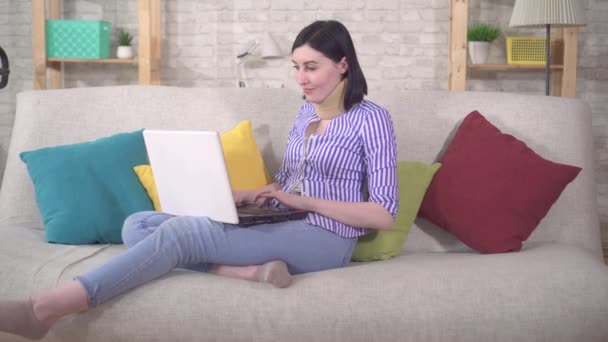 Joven mujer se sienta en el sofá con un collar alrededor de su cuello y utiliza un ordenador portátil — Vídeos de Stock