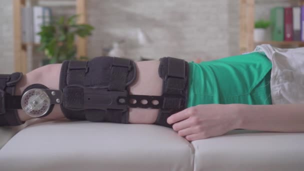 Pierna femenina con ortesis rodillera después de una lesión en el sofá médico — Vídeos de Stock