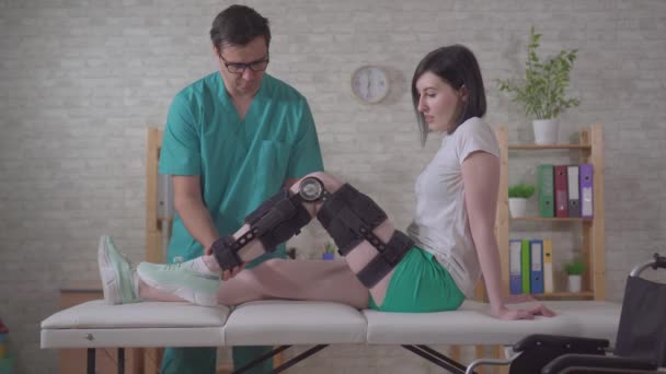 Fisioterapeuta masculino ayuda a dons ayuda con ortesis rodillera después de la lesión — Vídeos de Stock