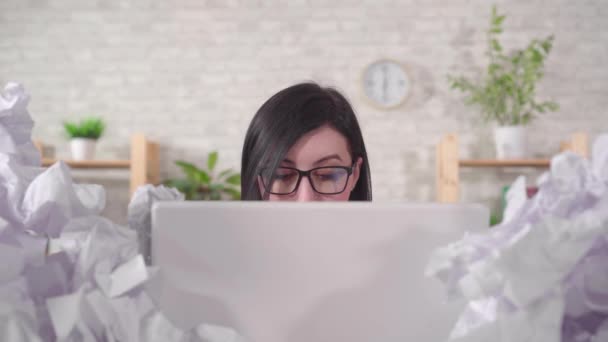 woman office worker with glasses is working at a laptop in a heaps of paper on an office desk