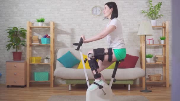 Jonge vrouw knie buigen orthose knie brace na letsel op een stationaire fiets knie buigen orthose knie brace na letsel — Stockvideo