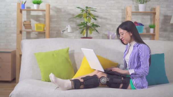 A portrait asian female with laptop after injury knee bend orthosis knee brace lies on the couch at home — 图库视频影像