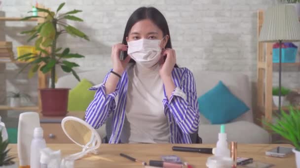 Joven mujer asiática se pone una máscara médica mientras se sienta en casa en una mesa y mira a la cámara — Vídeos de Stock