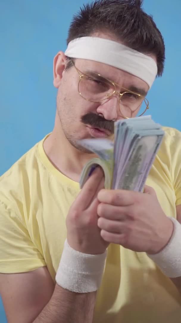 Retrato de un joven y divertido hombre bigotudo al estilo de los años 80 con un paquete de billetes sobre un fondo azul video vertical — Vídeos de Stock