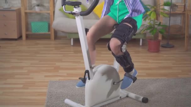 Um retrato mulher asiática joelho dobrar órtese joelho cinta após lesão em uma bicicleta estacionária joelho dobrar órtese joelho cinta após lesão — Vídeo de Stock