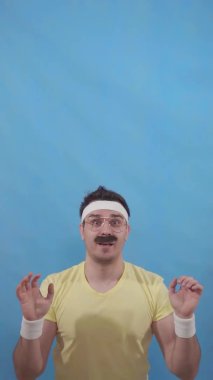 Mavi arka plan dikey videosunda 80 'lerin tarzında komik bıyıklı genç bir adam.