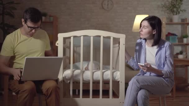 Young parents search the Internet and a newborn in a baby bed — ストック動画