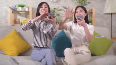 Two joyful young asian women scatter paper banknotes sitting on the sofa slow mo