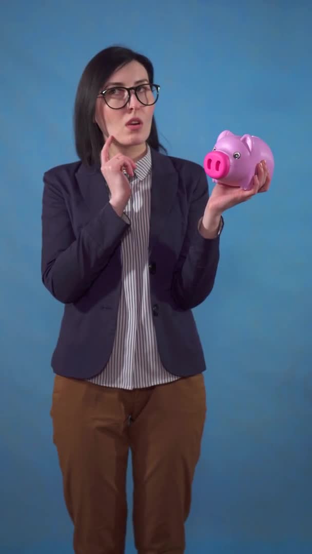 Молодая деловая женщина в костюме размышляет с копилкой в руках и находит решение на синем фоне вертикального видео — стоковое видео