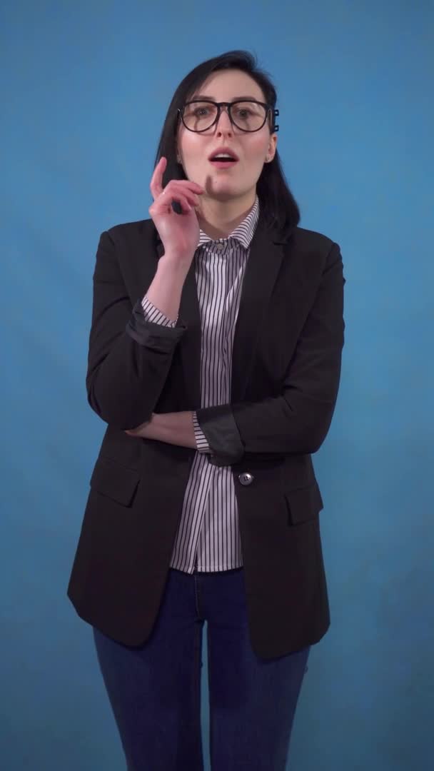 Thoughtful young business woman in a suit finds a solution standing on a blue background of a vertical video — Stock Video