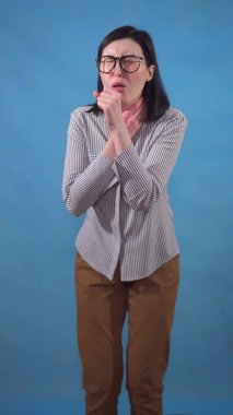 Genç bir kadın mavi arka planda dikey bir videoda dururken boğaz ağrısı ve öksürük yaşar.