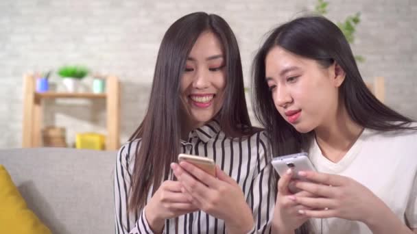 Dos jóvenes niñas asiáticas felices con teléfonos inteligentes en el sofá en la sala de estar de cerca — Vídeo de stock