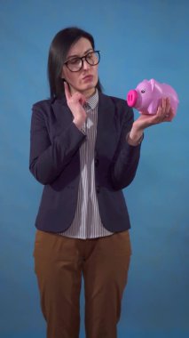 young business woman suit pondered with a piggy Bank in her hands on a blue background of a vertical video