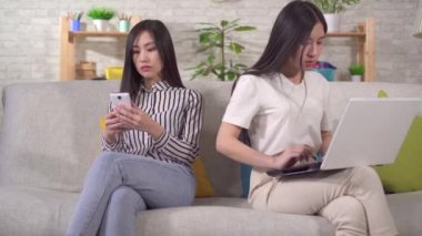 Two young asian women are using gadgets sitting on the sofa