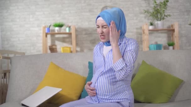 Young pregnant Muslim woman in a hijab will experience headaches sitting on the couch — Stock videók