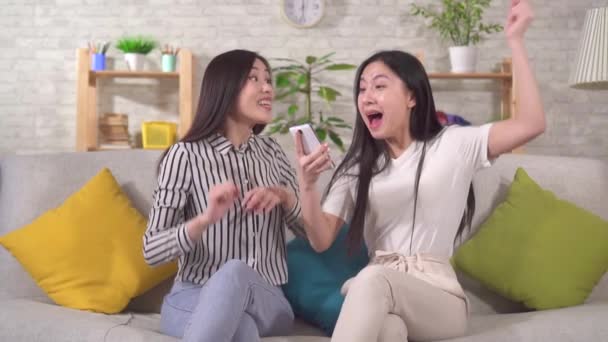 Two happy asian girls are sitting on the sofa in the living room happy to win looking at their smartphone slow mo — Wideo stockowe