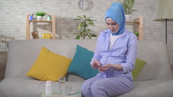 Мусульманская молодая беременная женщина принимает таблетки, сидя на диване — стоковое видео