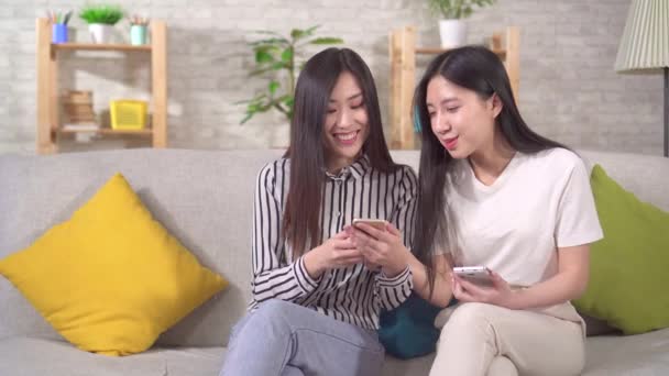 Két boldog fiatal ázsiai lány okostelefonnal a kanapén a nappaliban — Stock videók
