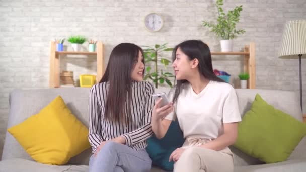 Two happy young asian woman use their smartphone sitting on the sofa — Stock Video