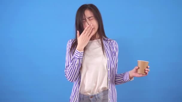 Junge schläfrige asiatische Frau gähnt und steht auf einem blauen Hintergrund mit einem nach oben von Kaffee — Stockvideo