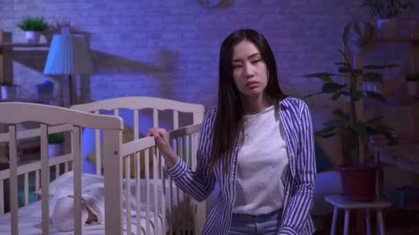Tired sad young asian mom rocks a baby cot — Stock Video