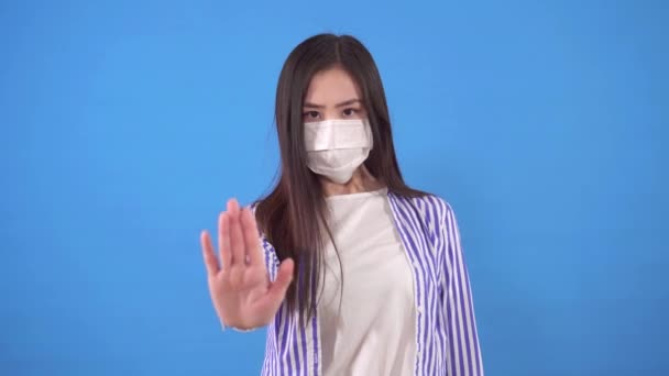 Молодая азиатская женщина в защитной медицинской маске на лице показывает стоп-жест, стоя на синем фоне — стоковое видео