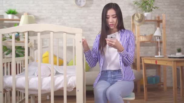 Молода азіатка зі смартфоном забиває дитину в дитячому ліжечку — стокове відео
