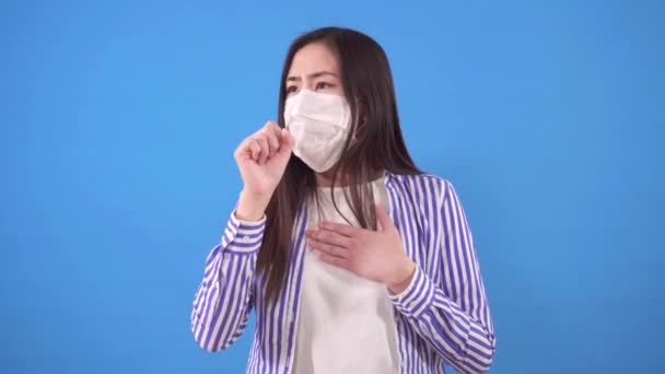 Krásná mladá asijská žena s ochrannou lékařskou maskou na tváři kašle, zatímco stojí na modrém pozadí — Stock video