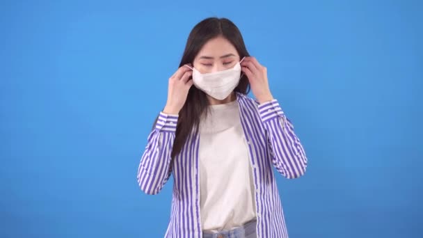 Schöne junge asiatische Frau setzt eine schützende medizinische Maske auf ihr Gesicht, das auf einem blauen Hintergrund steht — Stockvideo