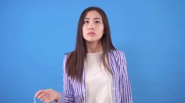 Genç ve çekici Asyalı bir kadın mavi bir arka planda duruyor. Yüzündeki koruyucu tıbbi maskeyi çıkarıyor.