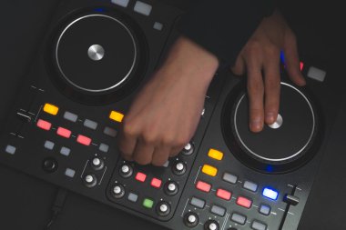 DJ müzik çalıyor. Ses karıştırıcı kontrolörü, düğmeleri ve sürgüleri var. Karanlığın içinde elleri ve pikapları aydınlatılmış kontrollerle karıştırın.