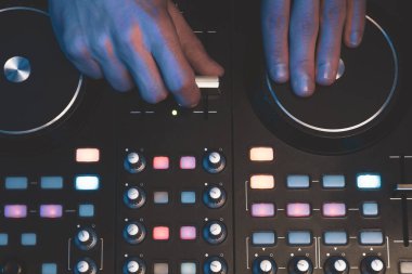 DJ müzik çalıyor. Ses karıştırıcı kontrolörü, düğmeleri ve sürgüleri var. Karanlığın içinde parlayan kontrollü pikaplarla karıştırma güvertesinde.