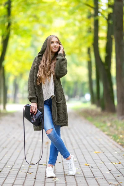 秋の公園でウォーキングと携帯電話を話すフード付きニット カーディガンを緑の美しい若い女性。秋のファッションのコンセプト. — ストック写真