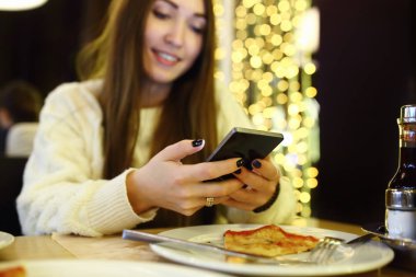 Kadın yazarak yazma iletide modern bir kafede akıllı telefon. Cep telefonu kullaranarak pizzayla bir masada oturan genç güzel kız Resim kırpılmış. Tonda. Seçici odak.