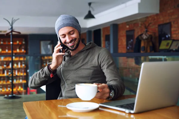 Junger gutaussehender Hipstermann mit Bart sitzt im Café und telefoniert, hält eine Tasse Kaffee in der Hand und lächelt. Laptop auf Holztisch. — Stockfoto