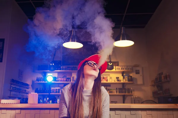 Mulher bonita nova na fumaça de boné vermelho um cigarro eletrônico na loja de vape — Fotografia de Stock