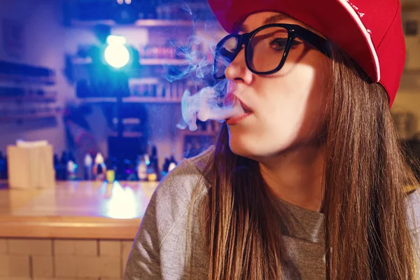 Wanita muda bertopi merah merokok rokok elektronik di toko vape. Penutup . — Stok Foto