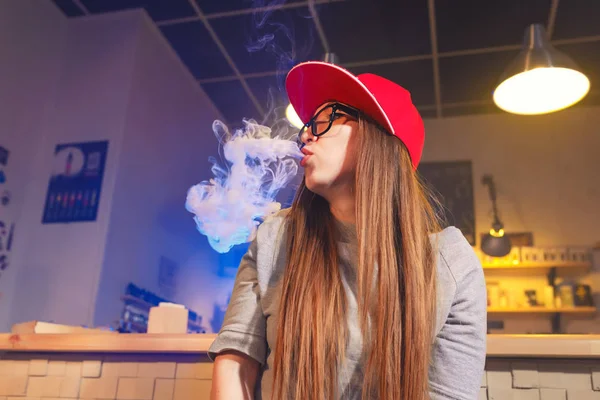 Mulher bonita nova na fumaça de boné vermelho um cigarro eletrônico na loja de vape — Fotografia de Stock