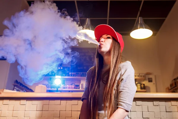 Jeune jolie femme en bonnet rouge fume une cigarette électronique à la boutique de vape — Photo